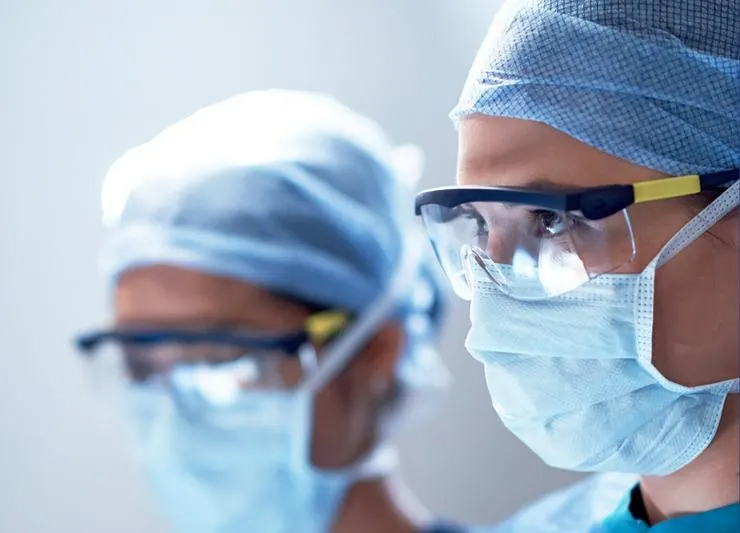 Two surgeons in scrubs, headscarves, safety goggles and surgical masks